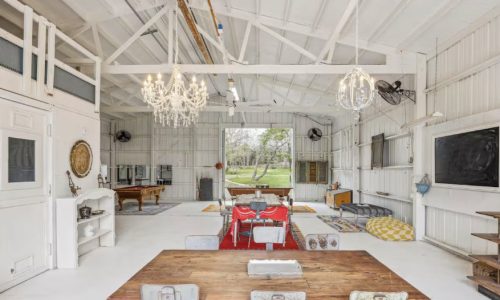 A spacious, open-plan entertaining area with a dining table, a pool table, and a loft space above. The space has white walls and ceiling with exposed beams, and colorful rugs on the floor.