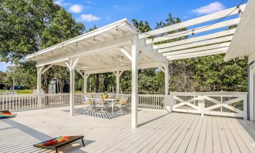 A white wooden deck with a covered pergola, outdoor dining set, and cornhole boards under a blue sky with trees in the background.