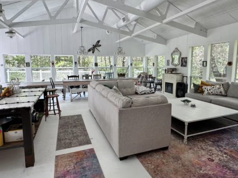A spacious, open-concept living and dining area with a large sectional sofa, a white coffee table, a dining table, a kitchen island, and large windows overlooking a wooded area.