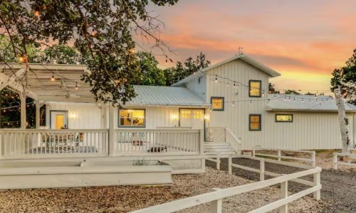A white farmhouse with a metal roof is illuminated by string lights during a vibrant sunset. A large deck with a white railing extends from the house, and a white fence surrounds the property.